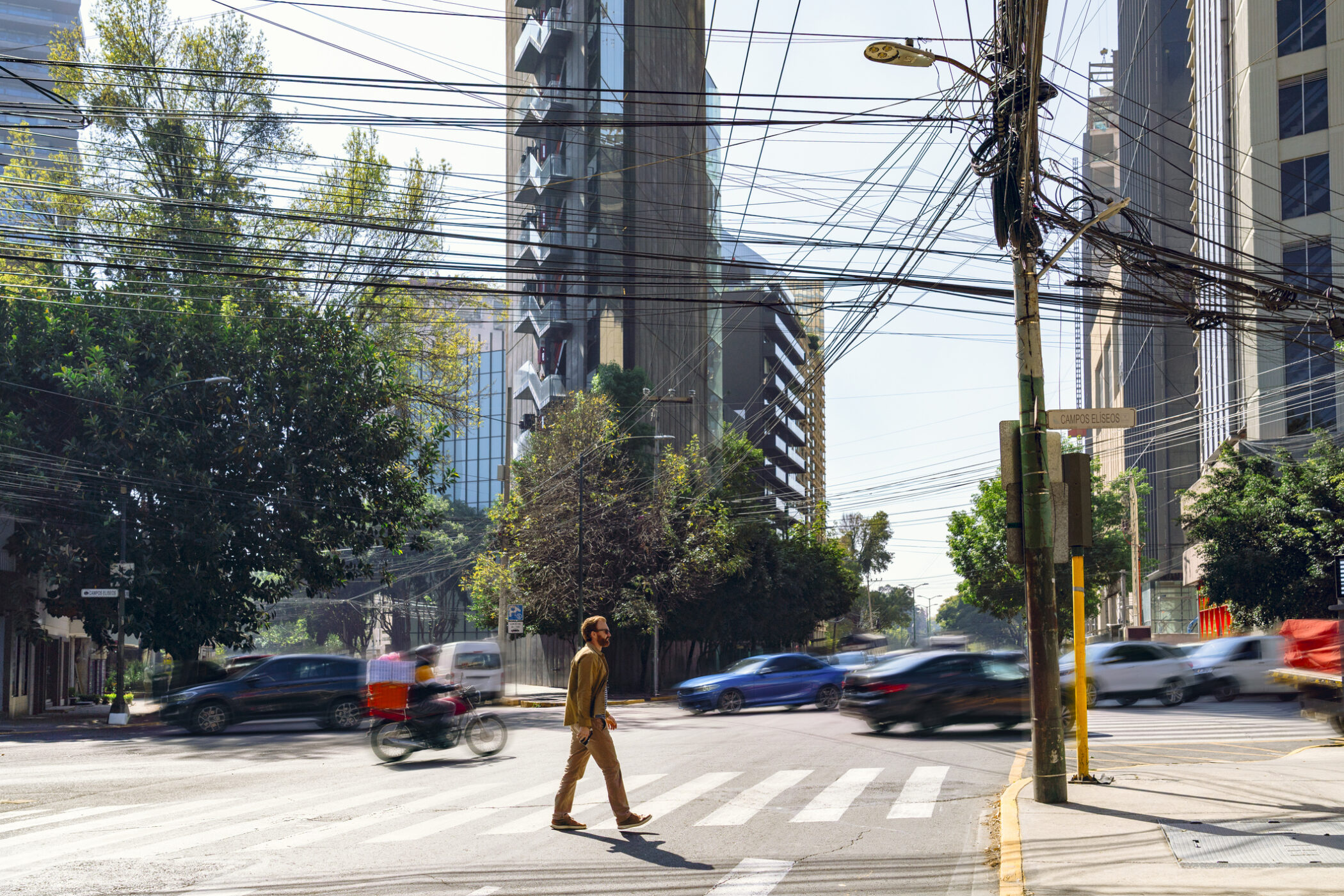 A man in casual attire crosses a busy intersection in Polanco, Miguel Hidalgo, Mexico City. Surrounding him are modern high-rise buildings, lush trees, and bustling traffic including cars and motorcycles. Overhead, numerous utility wires crisscross, adding to the urban landscape. The scene captures the dynamic and vibrant atmosphere typical of this affluent neighborhood.